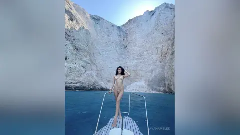 Fashionable woman in a gold bikini standing on a boat against a dramatic rocky cliff backdrop in the blue sea.