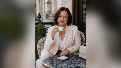 IsabellaBennet wears a white blazer and pearl necklace while sitting at a cafe table holding a white coffee cup.
