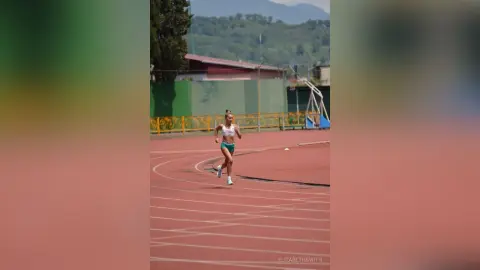 Athletic woman in white sports bra and teal shorts running on a red track at an outdoor stadium.