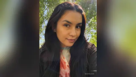 Smiling young woman with long dark hair wearing a patterned blouse in a sunny outdoor garden setting.