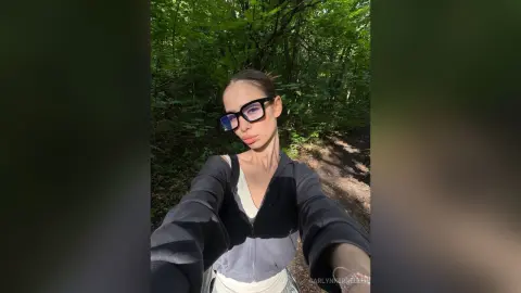 CarlynKernell wearing glasses and a dark zip-up jacket while posing outdoors in a sunlit, green forest setting.