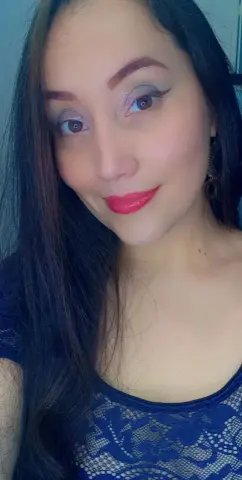 Smiling young woman with long dark hair and red lipstick wearing a blue lace top in a bright indoor setting.