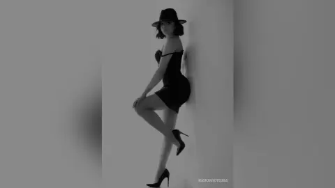 Elegant woman in a black cocktail dress and wide-brimmed hat posing against a minimalist wall in moody monochrome.