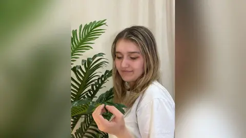 Young woman in a white t-shirt smiling while tending to a lush green indoor plant in a bright room.