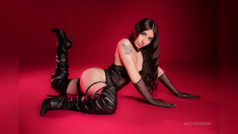 Glamorous woman in black corset and thigh-high boots posing against a vibrant red studio background.