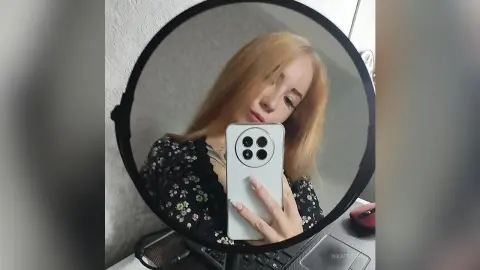 Blonde woman in a black floral blouse taking a selfie in a circular mirror with soft indoor lighting.
