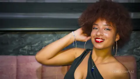 Smiling young woman with afro hair in a black halter dress posing in a moody lounge setting.