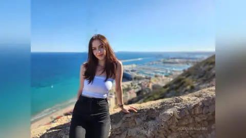 Smiling brunette woman in a white tank top and black jeans relaxing on a scenic coastal cliff overlooking the sea.