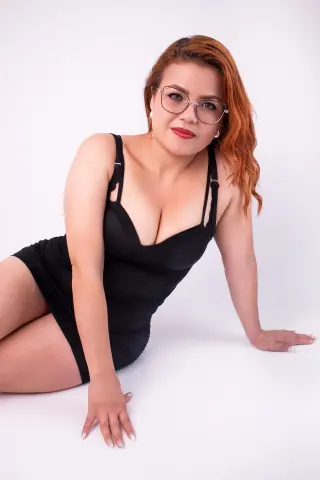 Red-haired woman in a black cocktail dress and glasses posing confidently against a white studio background.