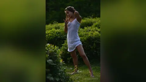 Dreamy young woman in a white summer dress posing in a lush green garden during golden hour.