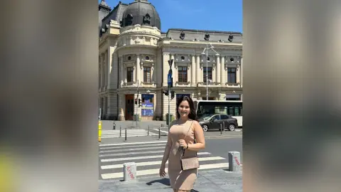 Smiling woman in a beige sleeveless dress standing in front of a grand European building on a sunny day.
