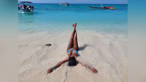 EnithAllegri lies on her back in the shallow ocean waves at a tropical beach, wearing a blue bikini top.