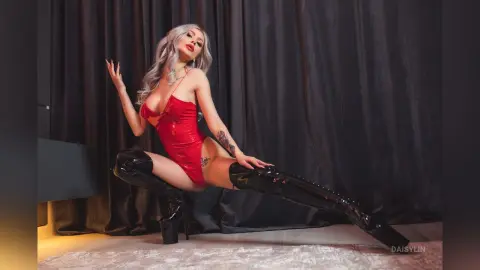 DaisyLin poses in a red shiny bodysuit and thigh-high black boots against a dark curtain backdrop.