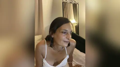 MichaelaBelle looks down while wearing a white lace camisole or bra top in a bedroom setting.