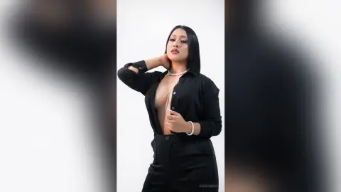 KimRoberth poses with one hand behind her neck, wearing a black button-down shirt unbuttoned to show her cleavage and a pearl necklace.
