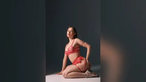 Elegant woman in red lace lingerie posing gracefully against a minimalist grey studio background.