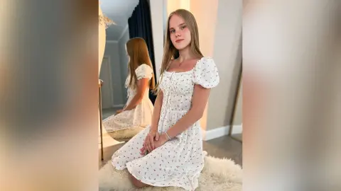 LiliLohan sits on a white furry rug wearing a white puff-sleeve floral print dress.