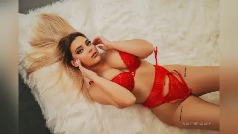 ValerieGracy lies on a white shag rug wearing red lace lingerie and showing her cleavage.