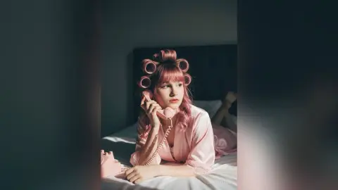 Young woman in pink silk pajamas with hair rollers using a vintage pink telephone in a moody bedroom setting.