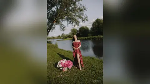 Elegant woman in a red floral maxi dress standing by a serene lake at sunset with pink flowers.