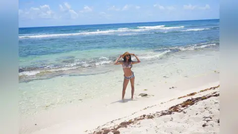 LucyMartin stands in shallow ocean water on a white sand beach wearing a patterned bikini and a sun hat.