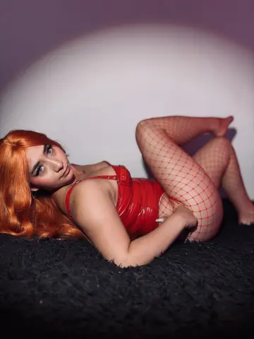 Redheaded woman in a red bodysuit and fishnet tights lying on a black shag rug in moody lighting.