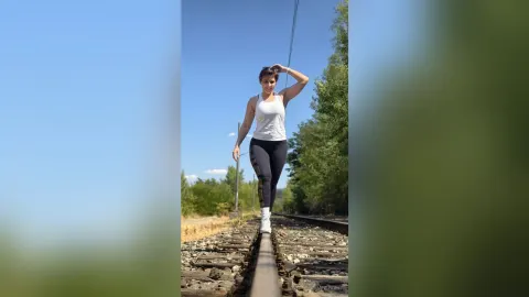 JaneHope — JaneHope walks along a railroad track wearing a white tank top and black leggings under a clear blue sky.