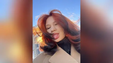 Smiling woman with flowing reddish hair wearing a black turtleneck and tan coat in bright sunlight.