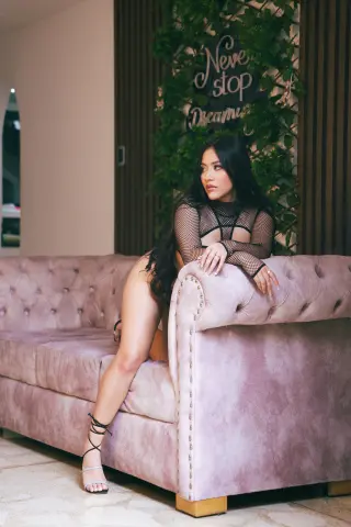 Nicol_Pierce sits on a pink velvet couch wearing a black mesh bodysuit and strappy high heels.