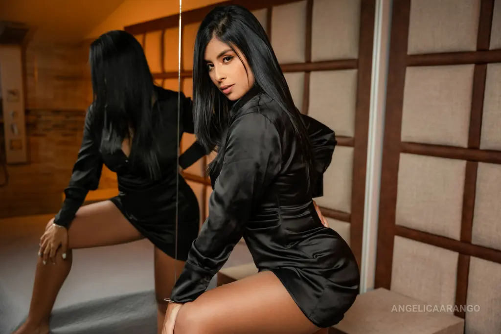 AngelicaArango poses naked in a black bodysuit, kneeling on a white block against a dark background.