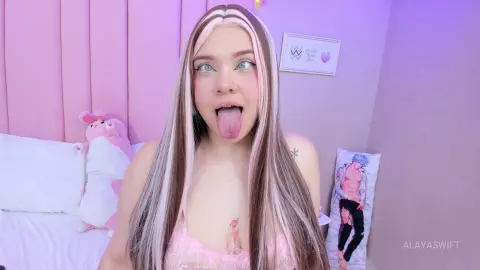 Young woman with long blonde hair and pink outfit sticking her tongue out in a cozy pink bedroom.