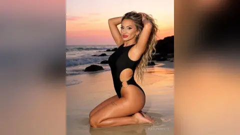 Blonde woman in a black cutout swimsuit posing on a sandy beach during a golden sunset.