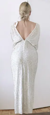 Blonde woman in a sparkling white sequin evening gown with a low back, standing in a bright minimalist room.