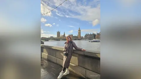 Stylish woman in a brown trench coat standing by the Thames river with London skyline in the background.