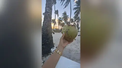 Person holding a fresh green coconut drink at a tropical beach resort during a sunny sunset.