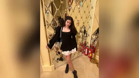 Young woman in a black long-sleeve dress posing elegantly in a luxury gold mirrored hallway.