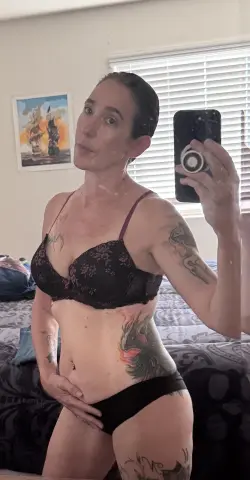 Tattooed woman in black lace lingerie taking a mirror selfie in a bright bedroom.