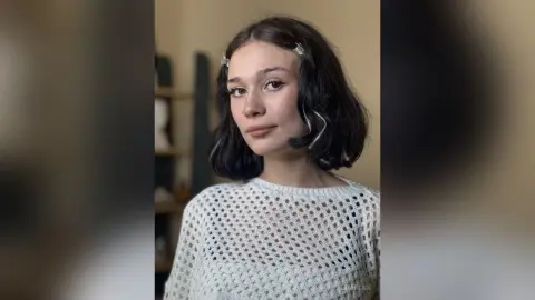 Young woman with wavy dark hair and hair clips wearing a white crochet knit sweater in a soft indoor setting.