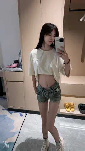 Young woman in a white oversized t-shirt and denim shorts taking a mirror selfie in a bright dressing room.