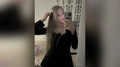 Young woman with long blonde hair in a black long-sleeve top taking a moody mirror selfie indoors.