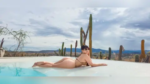 Woman in a bikini relaxing by a desert pool under a cloudy sky with cacti in a serene landscape.