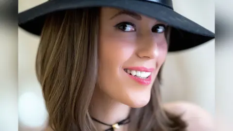 MontseFerrera smiles wearing a black wide-brimmed hat and a thin black choker necklace.