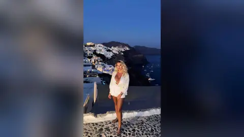 Blonde woman in a white summer outfit overlooking the coastal town of Santorini at twilight.