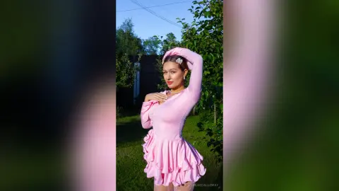 Young woman in a pink long-sleeve ruffled mini dress posing outdoors in a sunny garden setting.
