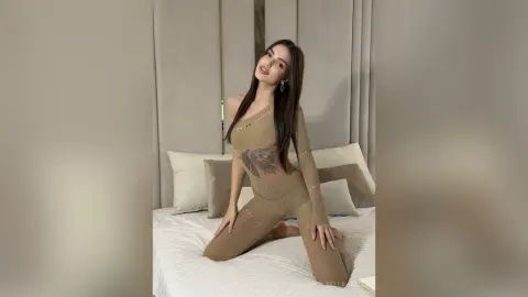 Elegant woman in a sheer beige bodysuit sitting on a white bed in a bright, minimalist bedroom.
