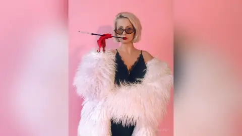 Glamorous woman in a white faux fur coat and black lace dress holding sunglasses against a pink background.
