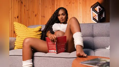 Brunette woman in a white crop top reading a book on a grey sofa against a warm wood paneled wall.