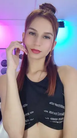 Young woman with red hair in a black tank top posing in soft pink and blue studio lighting.