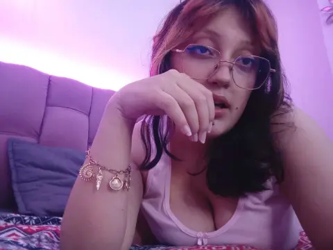 Magick_Black wearing glasses and a pink tank top, looking at the camera with her hand near her mouth in a bedroom setting.