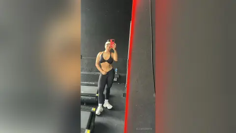 Fit woman in black sports bra and leggings taking a mirror selfie in a modern gym setting.